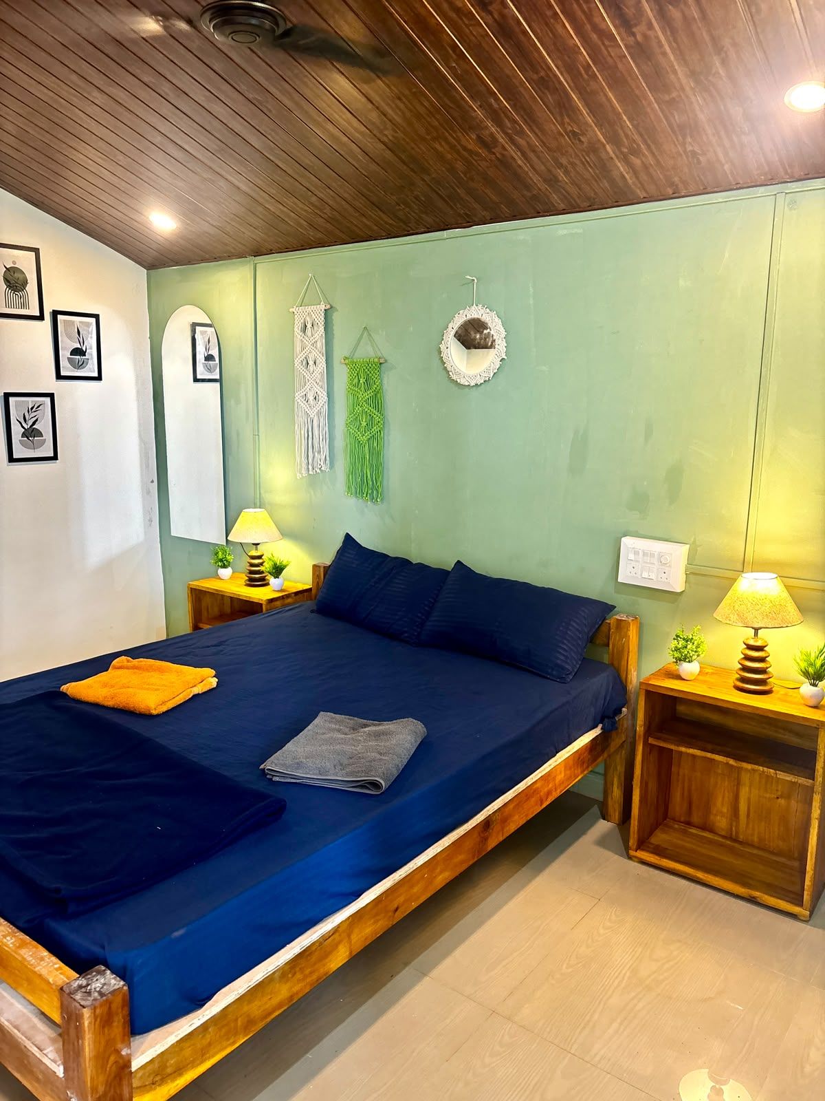 A bedroom with a wooden bed frame, a blue mattress, two pillows, a bedside table with a lamp, and a wall with framed artwork and macramé decorations.