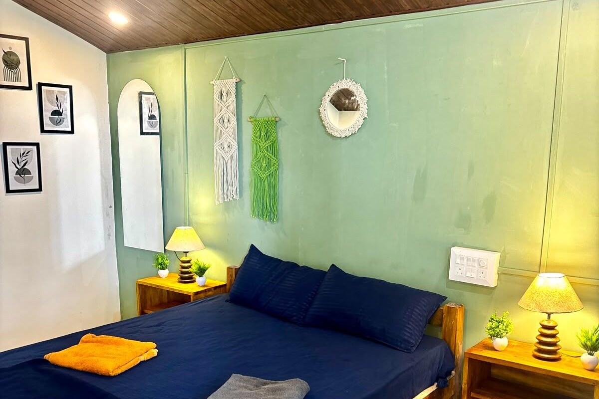 A bedroom with a wooden bed frame, a blue mattress, two pillows, a bedside table with a lamp, and a wall with framed artwork and macramé decorations.