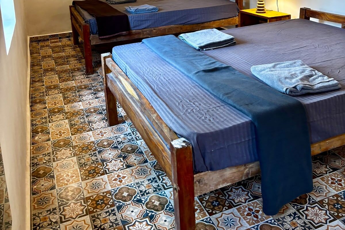 A room with two beds, a ceiling fan, a lamp, and patterned floor tiles.