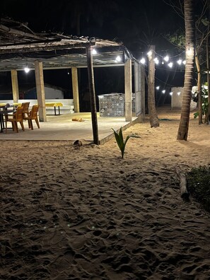 A covered outdoor area with a table and chairs, a small plant, and a sandy ground. - Absolute Paradise (Gokarna)