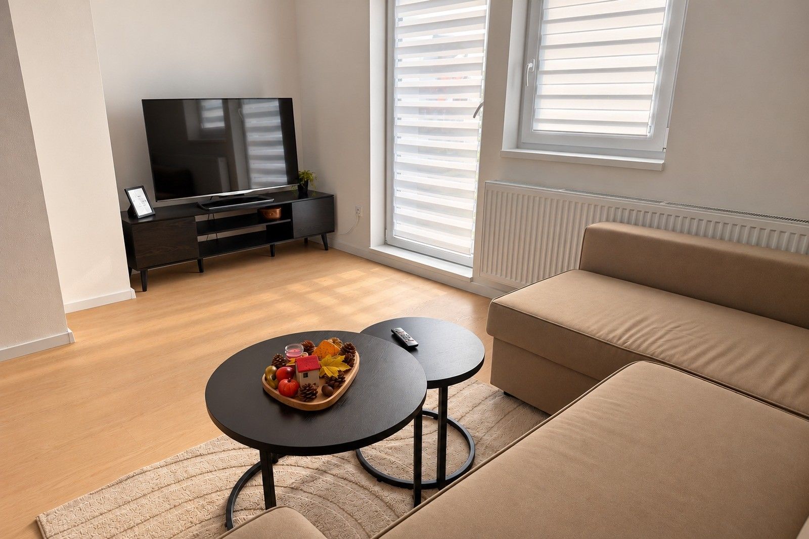 Ein modernes Wohnzimmer mit einer Couch, einem Couchtisch mit Obstschale, einem Fernseher auf einem schwarzen Lowboard und einem Fenster mit Jalousien.
