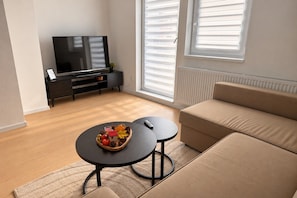 Ein modernes Wohnzimmer mit einer Couch, einem Couchtisch mit Obstschale, einem Fernseher auf einem schwarzen Lowboard und einem Fenster mit Jalousien.