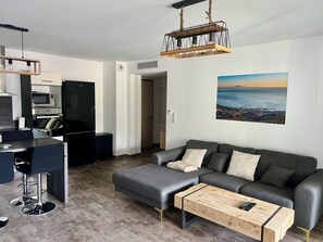 Een moderne woonruimte met een grijze bank, een houten salontafel, een keukenhoek met een magnetron, en een groot raam met uitzicht op de stad.