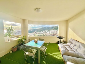 Een kamer met een balkon, een tafel met stoelen, een bank en uitzicht op een stadsbeeld.
