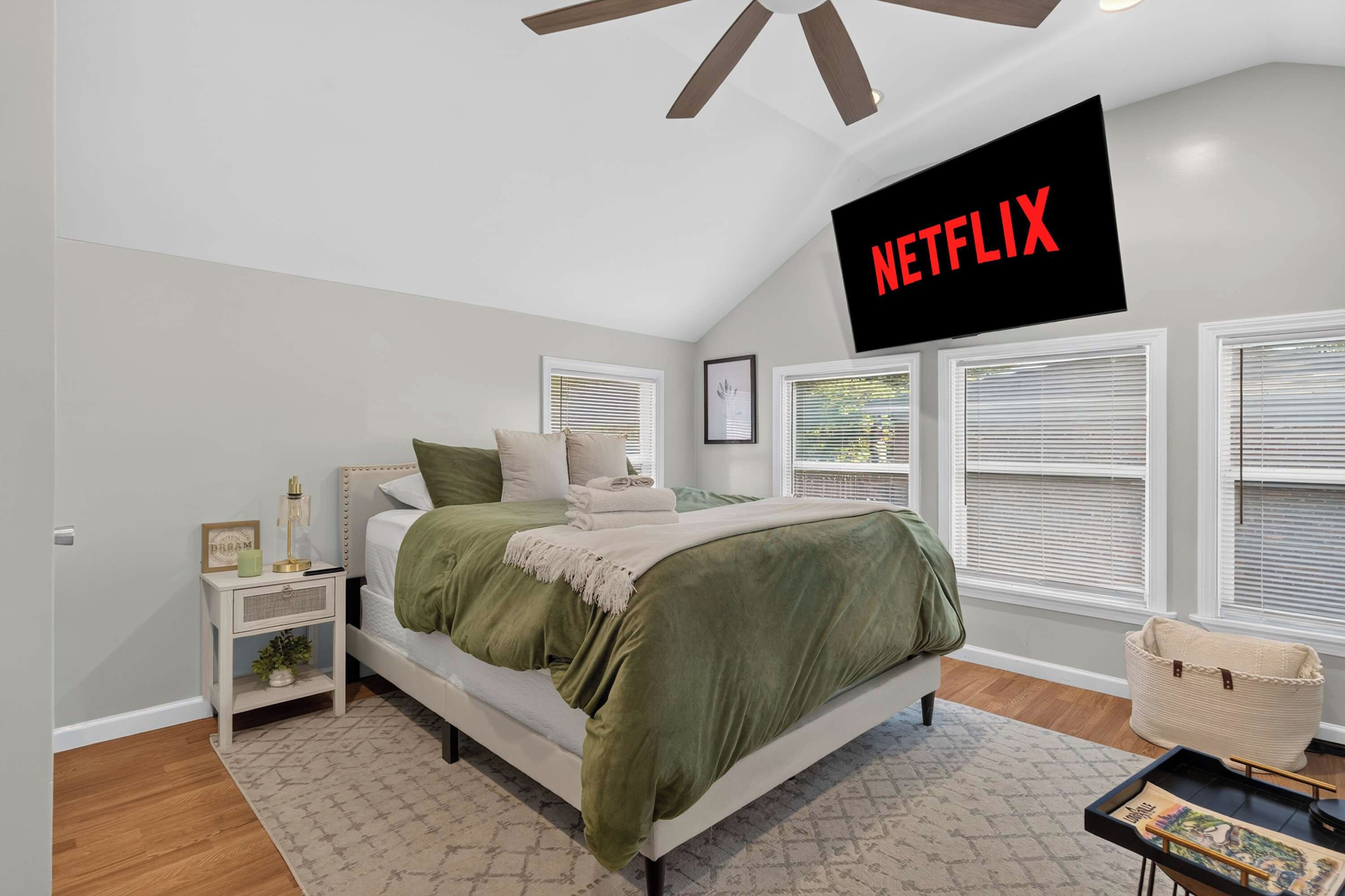 Une chambre à coucher comprenant un lit, des tables de chevet, un téléviseur fixé au mur et une fenêtre avec des stores.