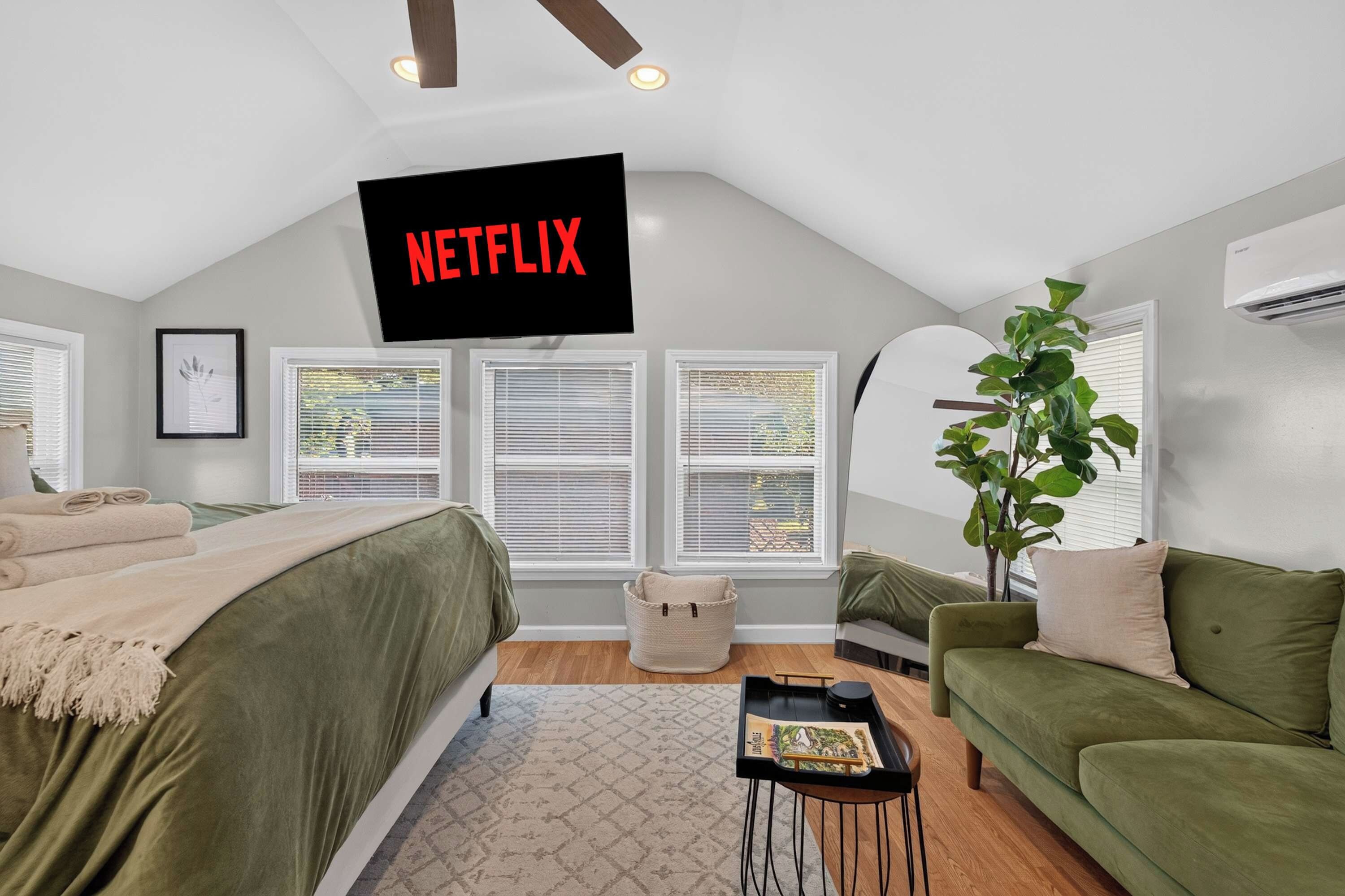 Une chambre avec un lit, un canapé vert, une plante, un téléviseur fixé au mur qui affiche le logo « NETFLIX », et une fenêtre avec des stores.