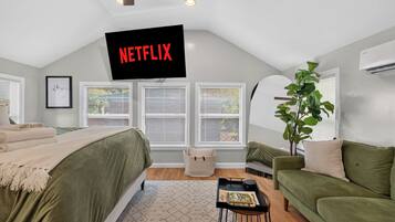 Une chambre avec un lit, un canapé vert, une plante, un téléviseur fixé au mur qui affiche le logo « NETFLIX », et une fenêtre avec des stores.
