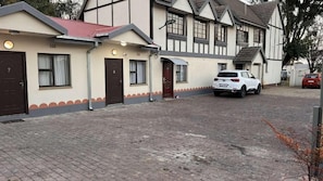 A two-story building with a brick driveway, a white car parked in front, and a red car in the background. - Los Angeles Gueshouse (Benoni)