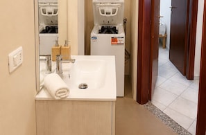 Une salle de bain avec un lavabo blanc, un miroir et une serviette. On aperçoit un distributeur d’eau et des toilettes à l’arrière-plan.