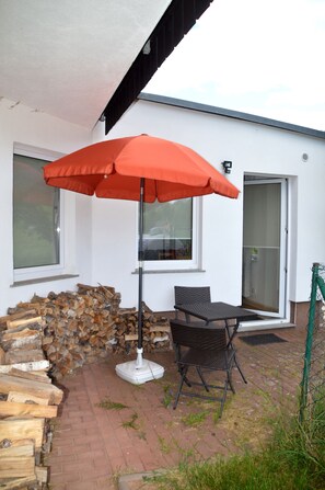 Ein Sitzbereich im Freien mit Tisch und Stühlen, einem roten Sonnenschirm und einem Holzstapel vor einem weißen Gebäude.