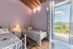 A bedroom with two beds, a nightstand with a lamp, and a view of a green landscape through a glass door. - Tenuta Bellosguardo (Toscana)