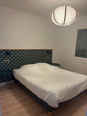 Une chambre à coucher comprenant un lit, une tête de lit, une table de chevet, une chaise, une fenêtre et un luminaire au plafond.