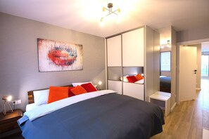 Ein modernes Schlafzimmer mit einem großen Bett, einem Nachttisch, einem Schrank und einem Kunstwerk an der Wand. Eine Tür führt in einen anderen Raum.