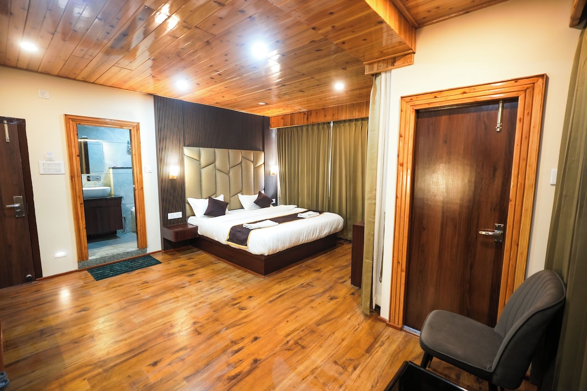 Een hotelkamer met een houten vloer, een bed met hoofdeinde, een nachtkastje, een stoel, een deur en uitzicht op de badkamer.