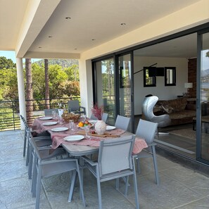Un espace de restauration en plein air avec une table dressée pour quatre personnes, entourée de chaises, et une vue sur l’intérieur à travers une porte vitrée.