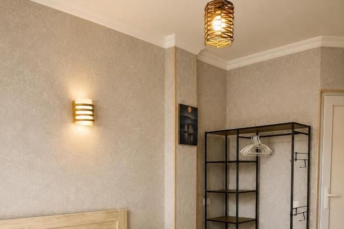 A hotel room with a bed, a wooden headboard, a wall-mounted shelf, a hanging light fixture, and a door.