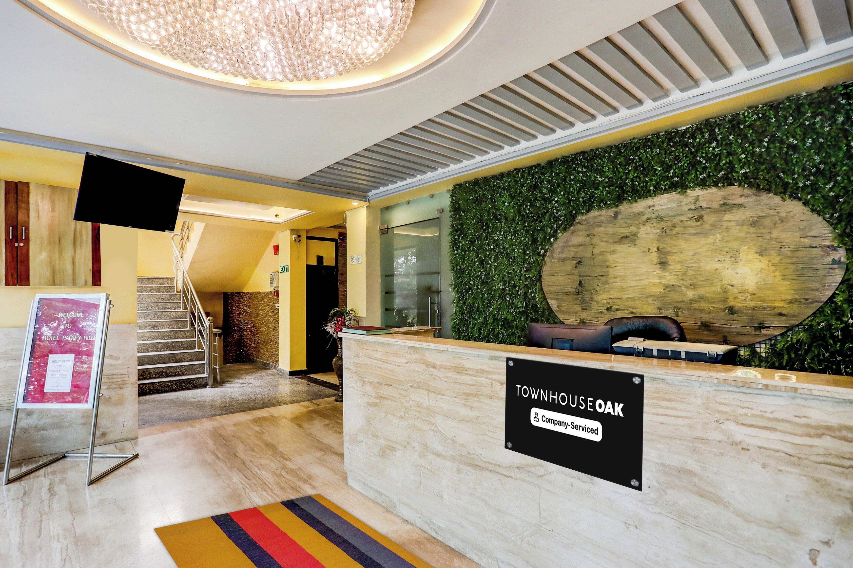 A hotel reception area with a modern design, including a reception desk, a staircase, and a sign indicating the property is serviced by a company.