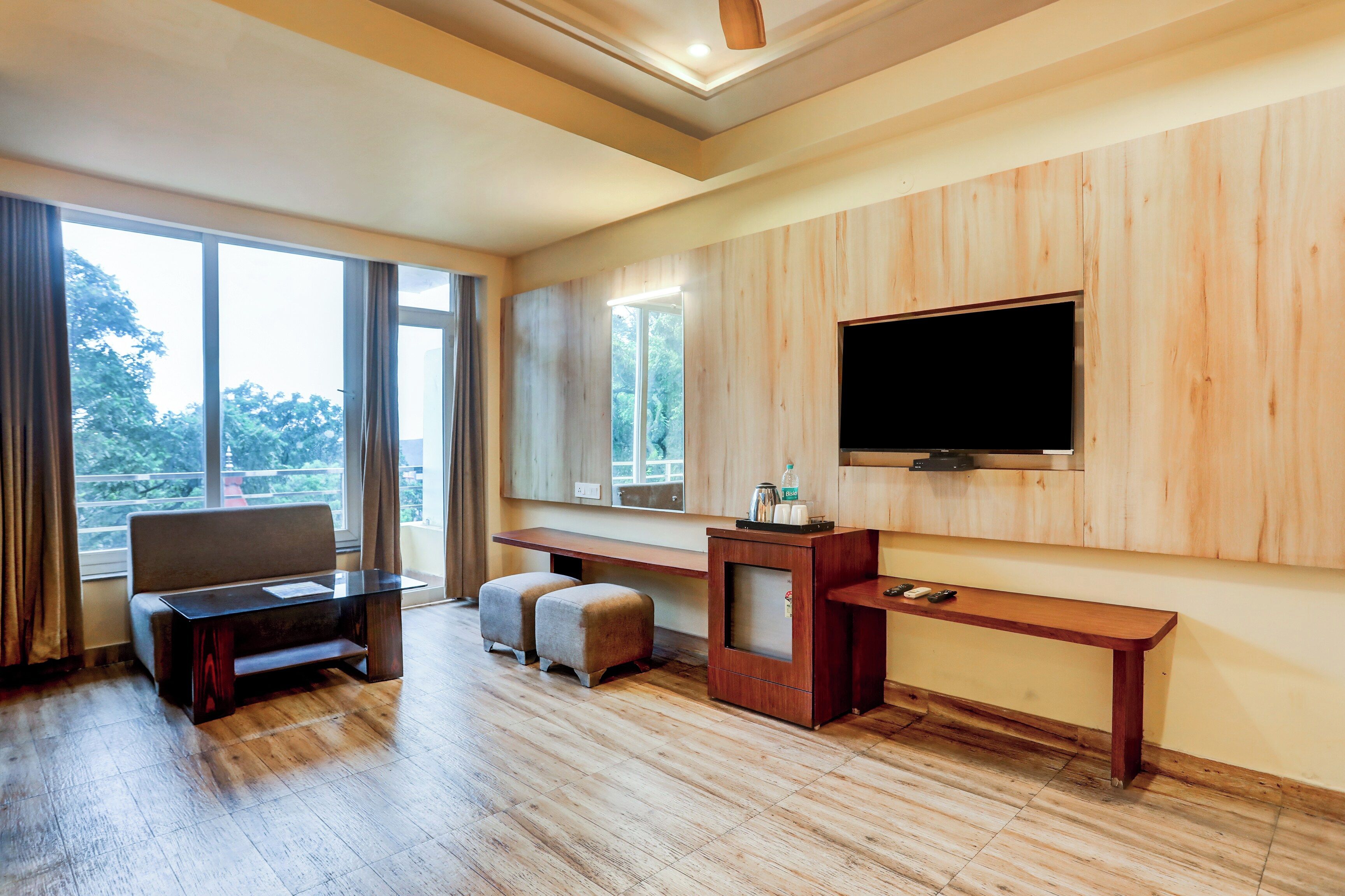 A hotel room with a large window, a flat-screen TV mounted on the wall, a wooden desk, a sofa, and a small ottoman.