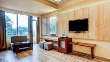A hotel room with a large window, a flat-screen TV mounted on the wall, a wooden desk, a sofa, and a small ottoman.