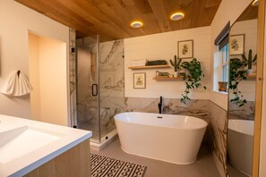 Ein modernes Badezimmer mit freistehender Badewanne, einer Duschkabine aus Glas und Holzregalen mit Pflanzen.