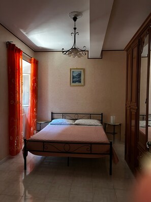 Ein Schlafzimmer mit Bett, Nachttisch, Stuhl, einem Bild an der Wand und einem Fenster mit Vorhängen.