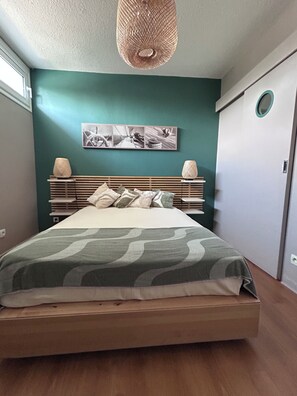 Une chambre à coucher avec un lit, une tête de lit, un tableau accroché au mur et une fenêtre ronde.