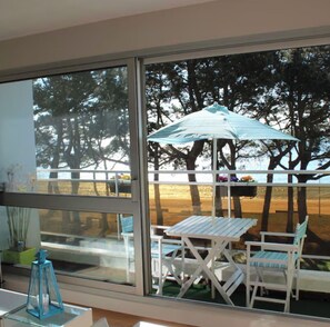 Un balcon avec une table et des chaises, un pot de fleurs suspendu, et une vue sur des arbres et une plage.