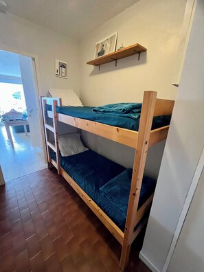 Een stapelbed met houten frame en blauw beddengoed, een klein plankje erboven en een deur naar een andere kamer.