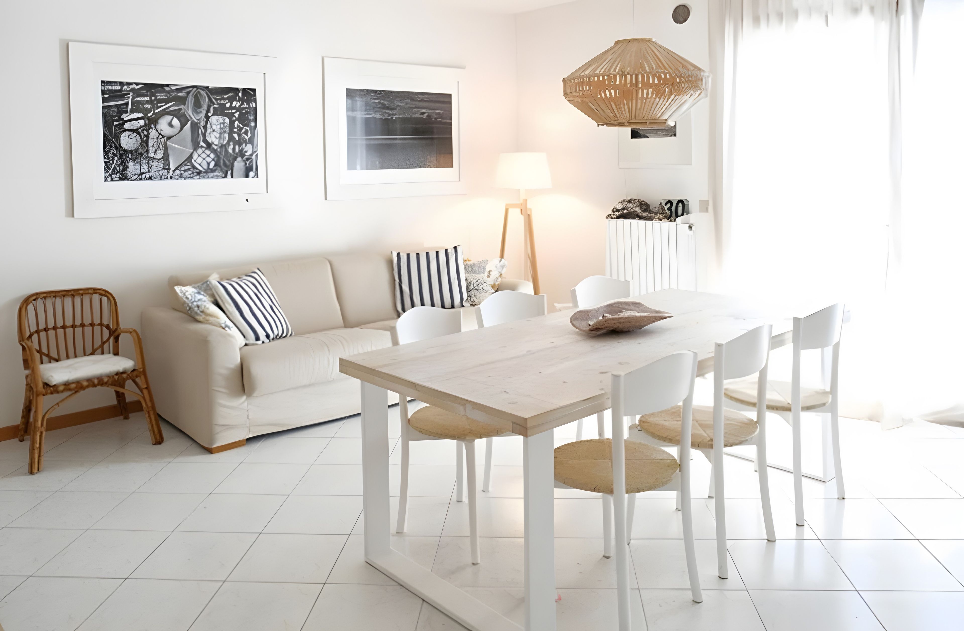 A modern living room with a white sofa, a dining table, and chairs. There are two framed pictures on the wall, a lamp, and a hanging light fixture.