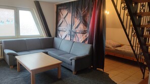 A room with a grey sofa, a wooden coffee table, a bed with patterned pillows, a window with blinds, and a staircase with a metal railing. - Apartment 'Marienmünster' with Wi-Fi (Marienmünster)