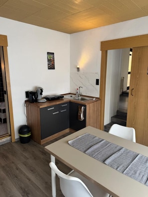 Cocina compacta con un lavamanos, una cafetera y un mueble pequeño. Hay una mesa de comedor con una silla y una alfombra.