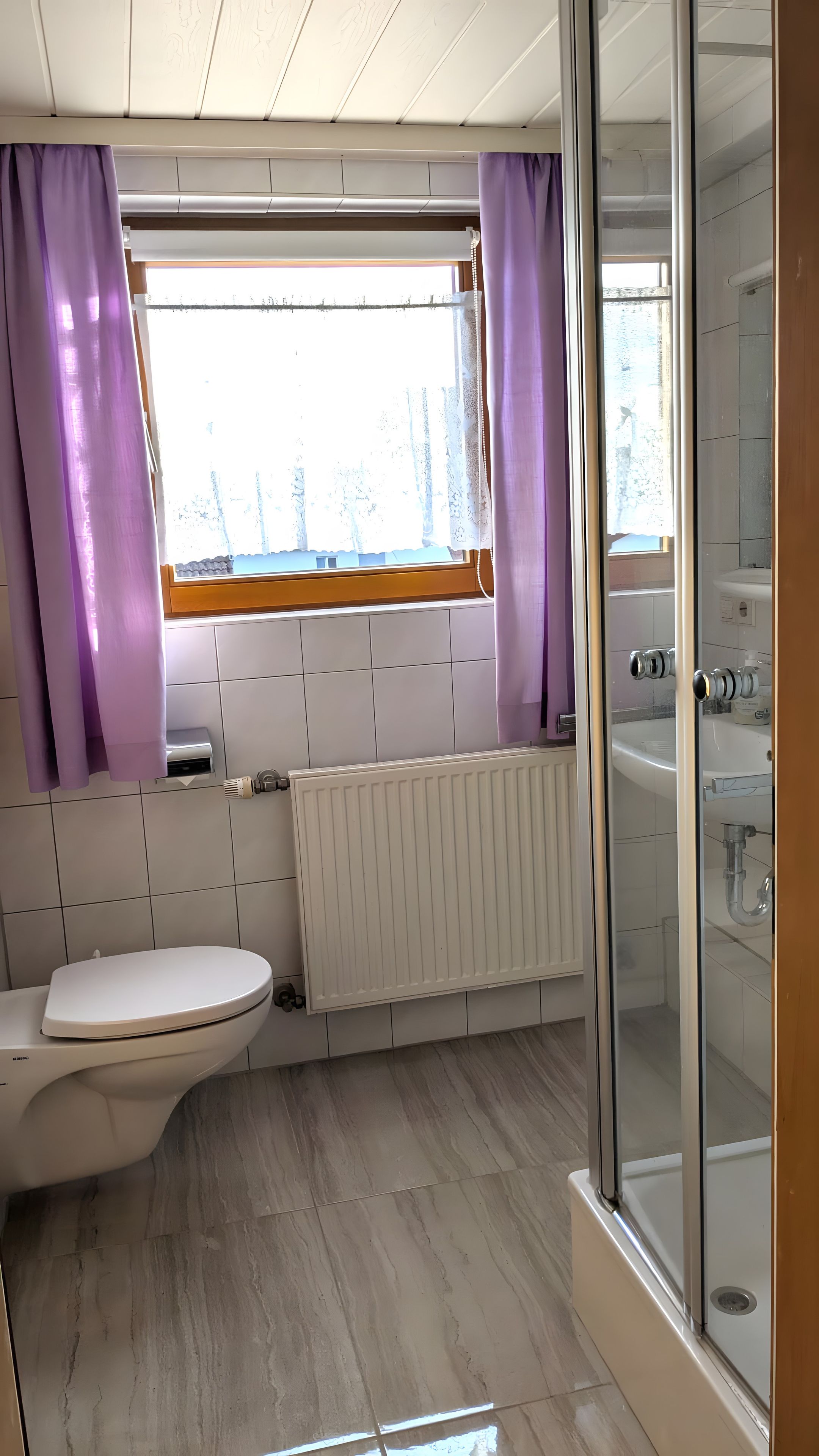 Ein Badezimmer mit Toilette, einem Fenster mit violetten Vorhängen, einem weißen Heizkörper und einer Duschkabine aus Glas.