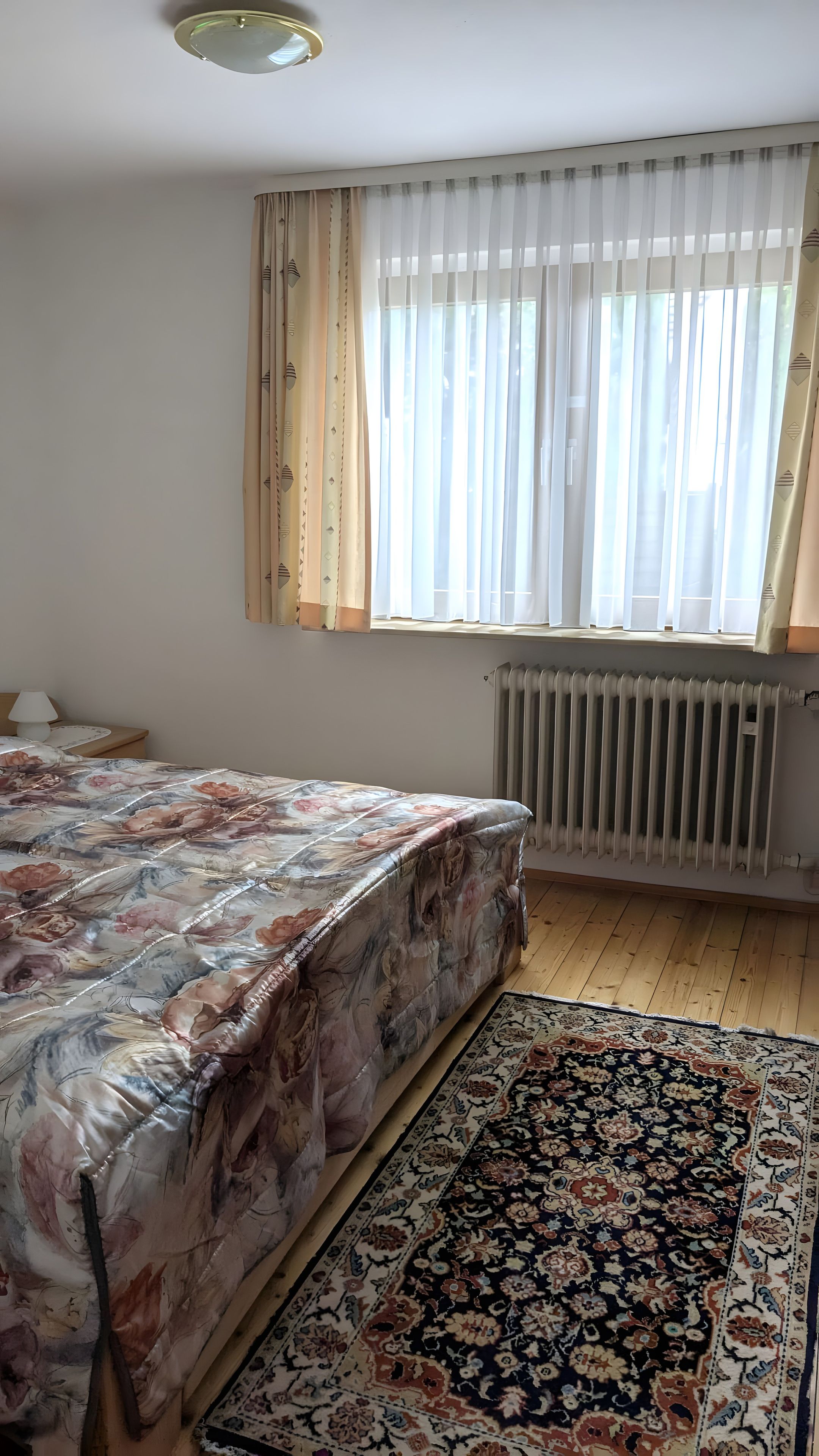 Ein Schlafzimmer mit einem Bett, einem Fenster mit Vorhängen, einem Heizkörper und einem Teppich.