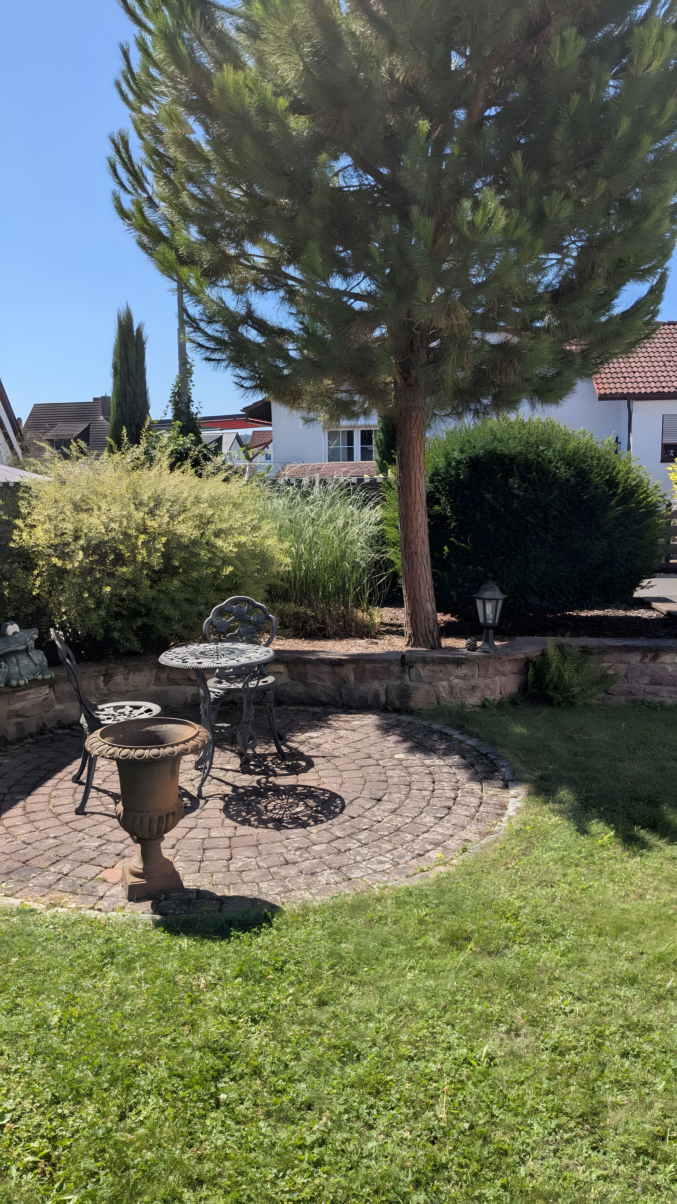 Ein Garten mit einer Terrasse, einem Tisch und Stühlen, umgeben von einer Steinmauer und verschiedenen Pflanzen.