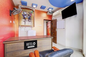 Een hotel receptiebalie met een 'HOTEL'-bordje, een aan de muur bevestigde televisie en een plafondventilator.