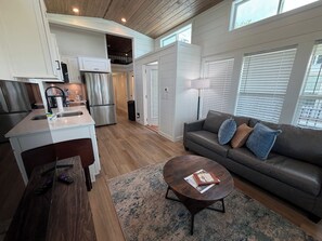 Una cocina moderna con refrigerador integrado, un comedor con mesa de centro redonda y una sala de estar con sofá y ventana con persianas.