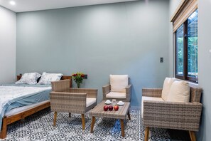 A bedroom with a bed, two wicker chairs, a small table with cups and apples, and a window with a view of greenery. - LEE NÔM VILLA DANANG (Ngũ Hành Sơn)