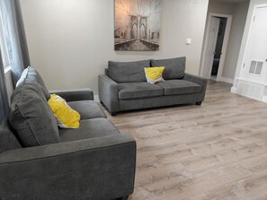 Uma sala de estar com dois sofás cinzas, um deles com almofada amarela, e um quadro na parede.