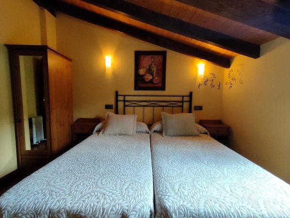Uma cama de casal com colcha estampada, cabeceira de madeira, duas mesas de cabeceira, um quadro na parede e um vigário de madeira no teto.