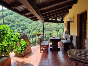 Um alpendre coberto com mesa e cadeiras, um vaso de pedra e vista para uma encosta arborizada.