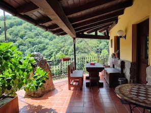 Um alpendre coberto com mesa e cadeiras, um vaso de pedra e vista para uma encosta arborizada.