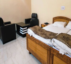 Quarto de hotel com cama de madeira, mesa de cabeceira, uma cadeira e pequena escrivaninha com gavetas.