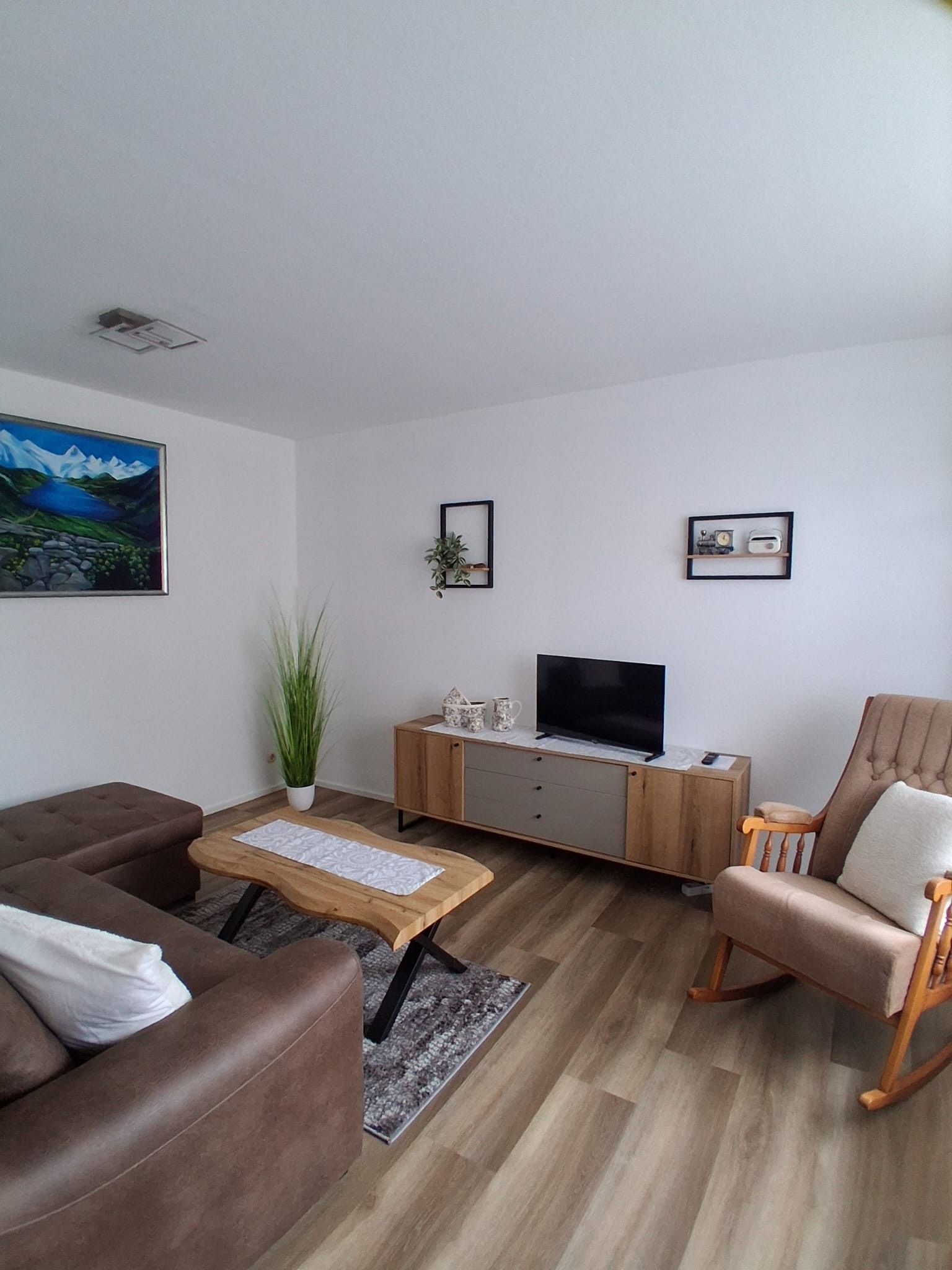 Ein modernes Wohnzimmer mit einem braunen Sofa, einem Couchtisch aus Holz, einem Fernseher auf einer Holzkommode und einem Schaukelstuhl.