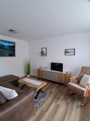 Ein modernes Wohnzimmer mit einem braunen Sofa, einem Couchtisch aus Holz, einem Fernseher auf einer Holzkommode und einem Schaukelstuhl.