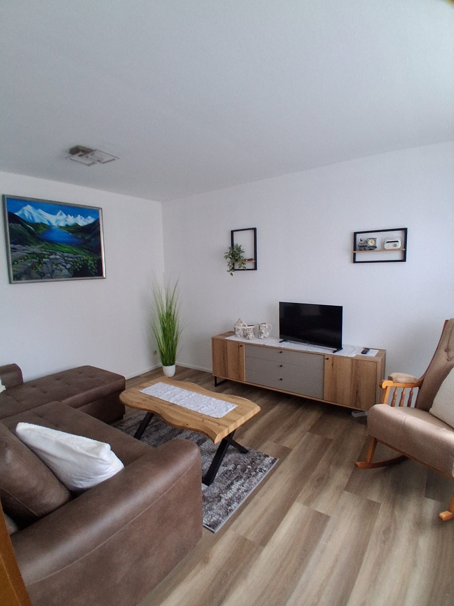 Ein Wohnzimmer mit einem braunen Sofa, einem Holztisch, einem Fernseher auf einem Schrank und einer Pflanze.