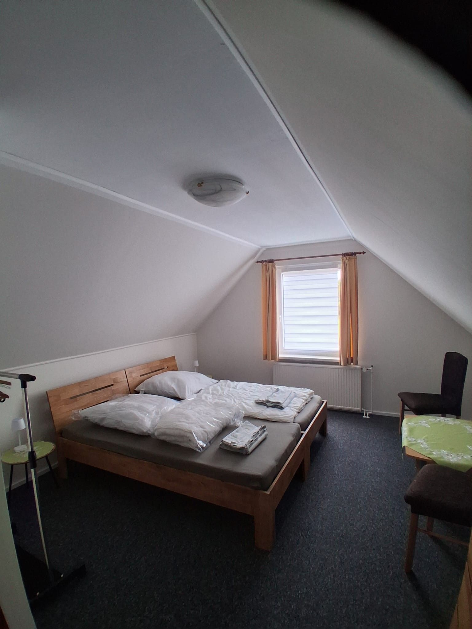 Ein kleines, schlichtes Zimmer mit einem Bett, einem Stuhl, einem Tisch und einem Fenster mit Vorhängen.