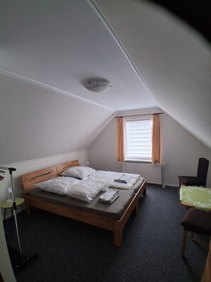 Ein kleines, schlichtes Zimmer mit einem Bett, einem Stuhl, einem Tisch und einem Fenster mit Vorhängen.
