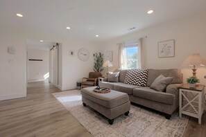 Una sala de estar con sofá, reposapiés y una mesita auxiliar, una ventana con cortinas y una puerta que conduce a otra habitación.