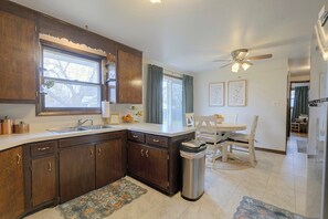 A kitchen with dark wooden cabinets, a white countertop, a sink, a ceiling fan, and a dining area with a table and chairs. - Walk to Lambeau! Cozy Green Bay Cottage! (Green Bay)