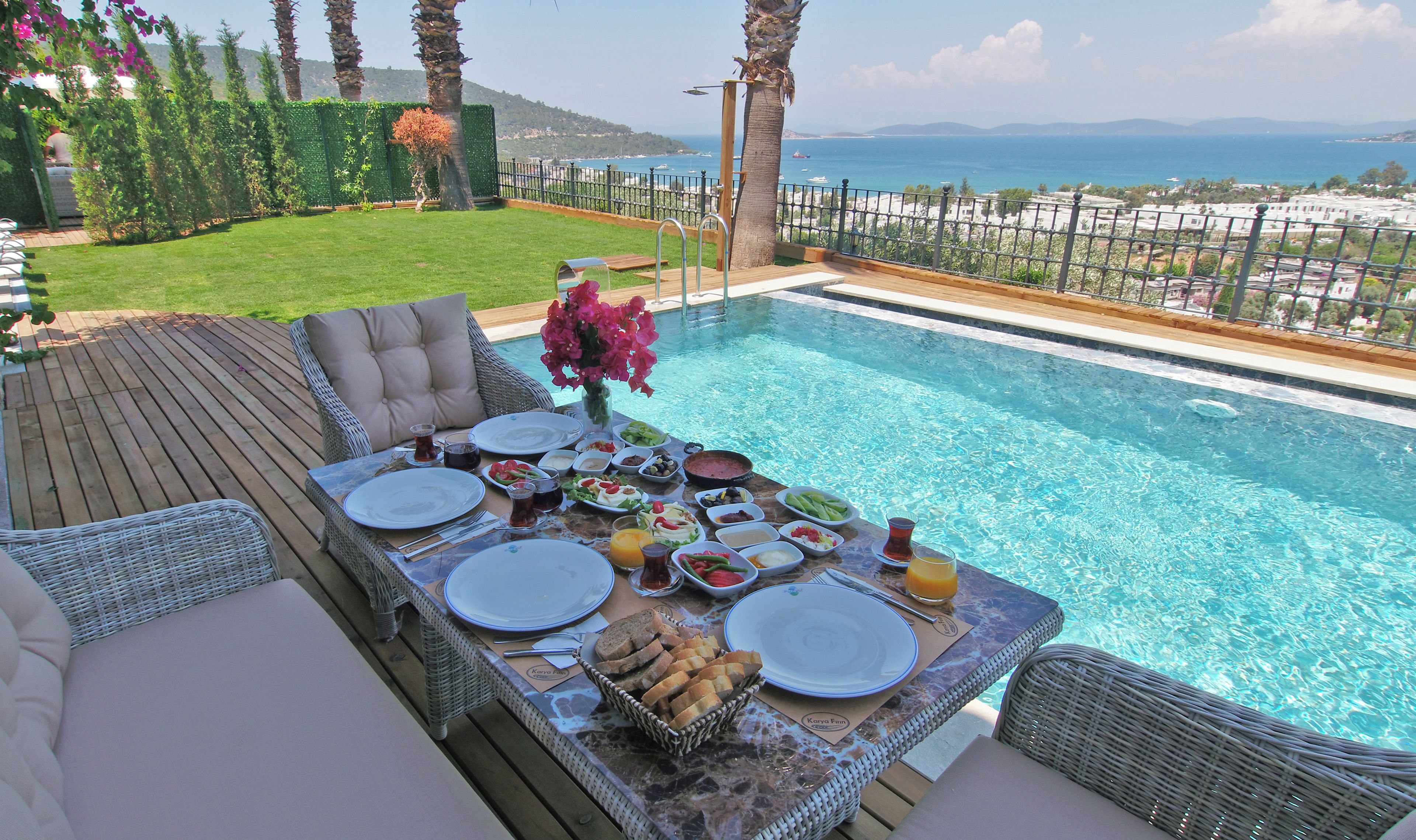 A poolside dining area with a table set for a meal, overlooking a pool and a coastal view.